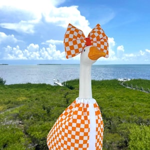 May include: A white goose figurine wearing a matching orange and white checkered fabric outfit and a large orange and white checkered bow on its head. The goose has a bright orange beak and orange feet, standing upright with a smooth, solid body.