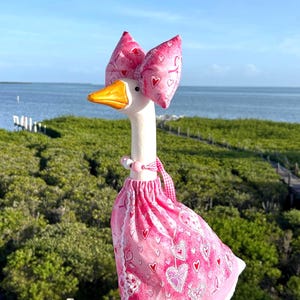 May include: A white goose figurine with an orange beak and feet, wearing a pink dress and a matching bow on its head. The fabric features a pattern of pink and white hearts. A necklace with pink and white beads is around the goose's neck.