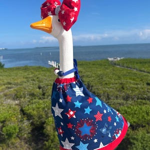 May include: A white ceramic goose figurine wearing a red fabric bow with white stars on its head and a blue fabric dress featuring a pattern of red, white, and blue stars and fireworks. The goose has an orange beak and orange feet on a green base.
