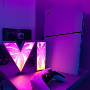Peut inclure: Une console PlayStation 5 blanche et une manette sans fil assortie placées à côté d'un panneau lumineux en forme de V. Le panneau présente une lumière dégradée rose et orange vive avec un motif de silhouette de palmier, diffusant un éclat vibrant.