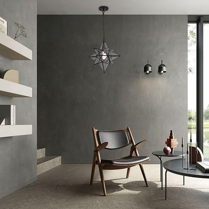 May include: A black metal star-shaped pendant light fixture hangs from the ceiling in a modern living room. The light fixture is surrounded by a gray wall, a black leather chair, and a wooden coffee table.
