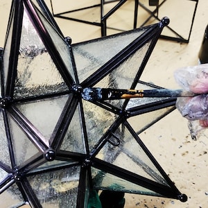 May include: A black metal star-shaped lantern with clear glass panels is being painted with a black paintbrush. The lantern is being painted on a light brown surface.