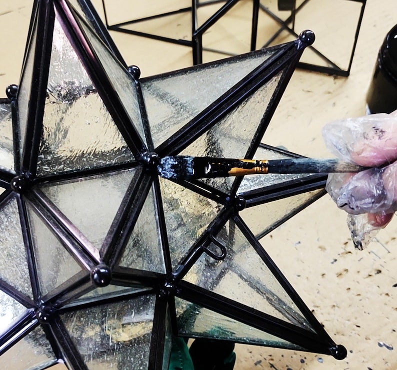 May include: A black metal star-shaped lantern with clear glass panels is being painted with a black paintbrush. The lantern is being painted on a light brown surface.