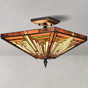 Può includere: Una plafoniera quadrata con paralume in vetro colorato con motivi geometrici nei toni dell'ambra, dell'arancione e del verde. L'apparecchio presenta una struttura in metallo color bronzo scuro, una base quadrata e un terminale decorativo.