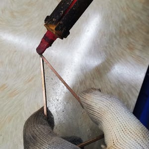 May include: A gloved hand holds a triangular piece of metal while a red soldering iron is used to melt solder onto the metal.