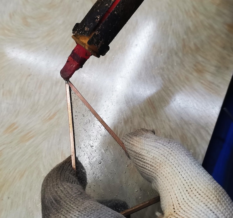 May include: A gloved hand holds a triangular piece of metal while a red soldering iron is used to melt solder onto the metal.