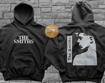 Może przedstawiać: Czarna bluza z kapturem z białym nadrukiem graficznym zespołu "The Smiths" z przodu i białym nadrukiem graficznym twarzy osoby z tekstem "The Smiths" z tyłu.