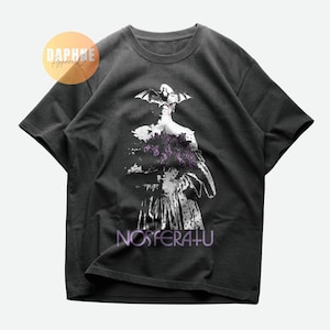 May include: A dark gray t-shirt with a white graphic print of a person with bat wings holding a bouquet of flowers. The text "NOSFERATU" is printed below the graphic.