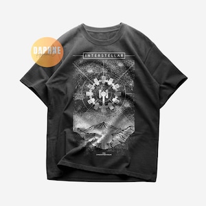 Könnte beinhalten: Dunkelgraues T-Shirt mit einer Grafik aus dem Film "Interstellar". Das Design zeigt eine Raumstation, eine Bergkette und den Filmtitel. Das T-Shirt enthält auch den Namen "Christopher Nolan."