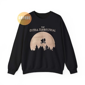 Puede incluir: Sudadera negra con una silueta de bicicleta y figura frente a una luna, sobre una línea de árboles. El texto "THE EXTRA-TERRESTRIAL" está impreso sobre la luna. Un logotipo de Daphne Apparel Co. está en la esquina superior izquierda.