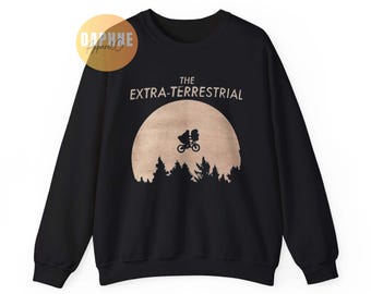Könnte beinhalten: Schwarzes Sweatshirt mit einer Silhouette eines Fahrrads und einer Figur vor einem Mond, über einer Baumlinie. Der Text "THE EXTRA-TERRESTRIAL" ist über dem Mond aufgedruckt. Ein Daphne Apparel Co. Logo befindet sich in der oberen linken Ecke.