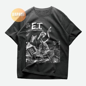 Könnte beinhalten: Dunkelgraues T-Shirt mit einer Schwarz-Weiß-Grafik von E.T. Der Außerirdische. Das Design zeigt E.T., der nach einem Stern greift, mit dem Text "E.T. The Extra-Terrestrial" und anderen Objekten.