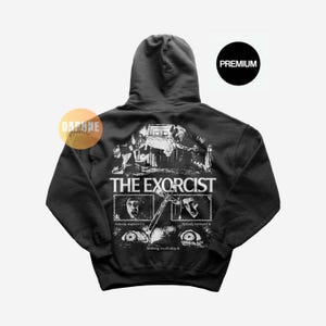 May include: A black hooded sweatshirt featuring a white graphic print on the back. The design includes the text THE EXORCIST, scenes from the film, a crucifix, and the phrases Nobody expected it, Nobody believed it, and Nothing could stop it.