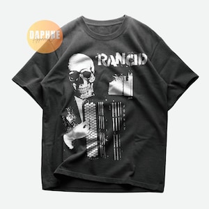 Peut inclure: Un t-shirt gris foncé avec un motif blanc d'un squelette jouant de l'accordéon. Le texte "Rancid" est imprimé au-dessus du squelette.