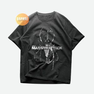 May include: Dark gray t-shirt featuring a white graphic design and the text "ANGEL MASSIVE ATTACK". The shirt has short sleeves and a crew neck. A logo is visible in the upper left corner.