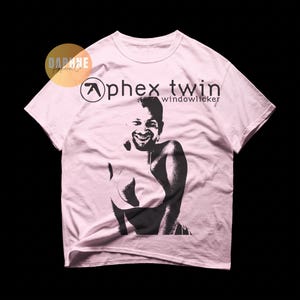 May include: A light pink short-sleeve t-shirt featuring a black graphic print of a smiling face on a body with the text Aphex Twin windowlicker and a circular logo. The shirt has a crew neckline and a standard fit with a soft cotton texture.