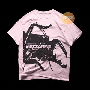 May include: A light pink short-sleeve t-shirt featuring a large black graphic of a beetle. The text "MASSIVE ATTACK" and "MEZZANINE" is printed in white over the center of the insect illustration. A small circular logo is visible in the top right.