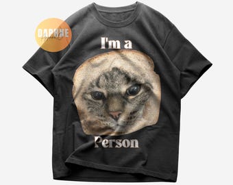 Puede incluir: Una camiseta negra de manga corta con un gráfico de la cara de un gato asomándose a través de una rebanada de pan. El texto "I'm a" aparece sobre el gato y "Person" debajo. Un logotipo circular dice "DAPHNE Apparel Co."