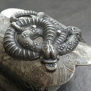 May include: A detailed, antique silver-toned container with a coiled snake design on the lid. The snake is intricately detailed, with textured scales and a detailed head. The container has ornate floral and scrollwork patterns.