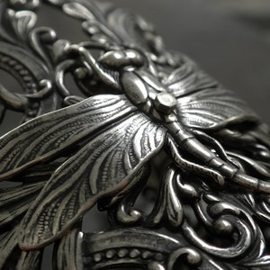 May include: Close-up of a silver-toned metal decorative piece featuring a dragonfly and ornate, swirling leaf and floral designs. The dragonfly is centered, with detailed wings and body. The piece has a vintage, antique aesthetic.