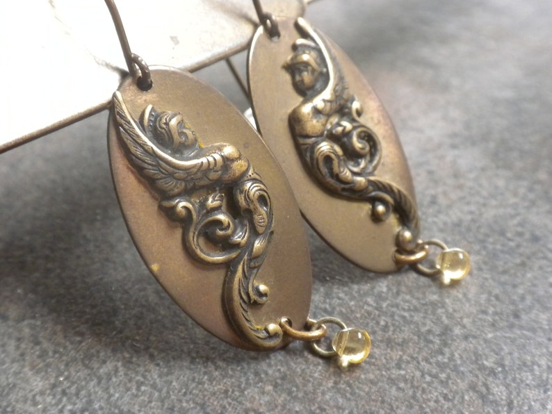 Puede incluir: Un par de pendientes de tono dorado con un dise&ntilde;o detallado y ornamentado que presenta un p&aacute;jaro y motivos florales. Cada pendiente est&aacute; acentuado con una peque&ntilde;a perla transparente.