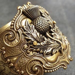 A brass cuff bracelet with floral etching and decorative layer features a Scottish thistle as the focal point.