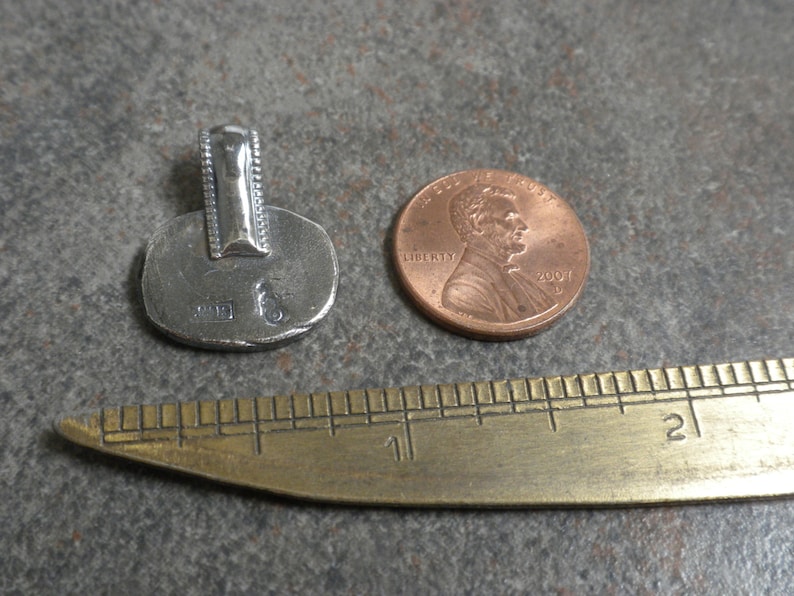 May include: Silver pendant with a textured surface and a bail. The pendant is about the size of a US penny.