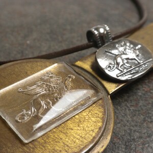 May include: A silver pendant necklace with a winged lion design. The pendant is on a brown leather cord and is displayed on a gold-colored surface.