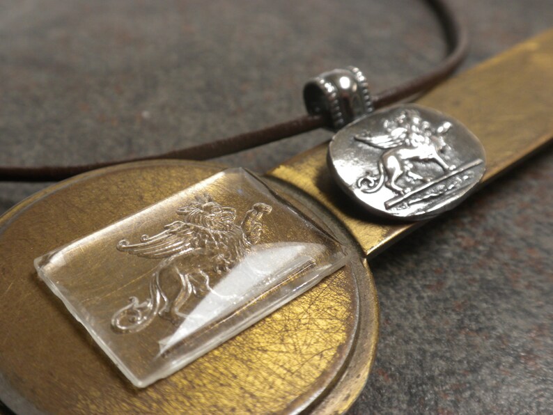 May include: A silver pendant necklace with a winged lion design. The pendant is on a brown leather cord and is displayed on a gold-colored surface.