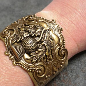 May include: Ornate gold-toned cuff bracelet featuring a detailed thistle design. The cuff has a textured surface with intricate scrollwork and a beaded edge. The thistle is the focal point, with detailed leaves and a textured seed head.