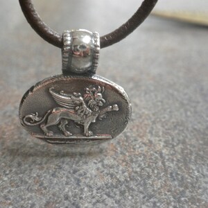 May include: A silver pendant necklace with a brown leather cord. The pendant features a detailed depiction of a winged lion.