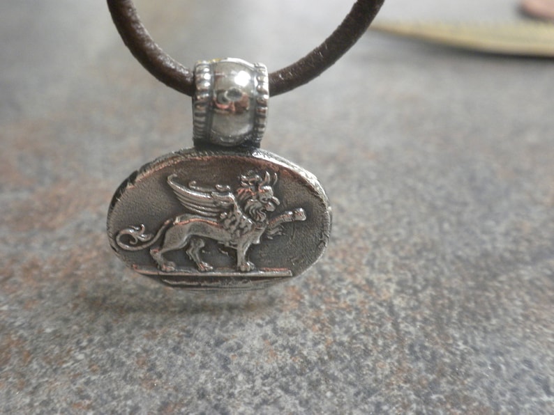 May include: A silver pendant necklace with a brown leather cord. The pendant features a detailed depiction of a winged lion.
