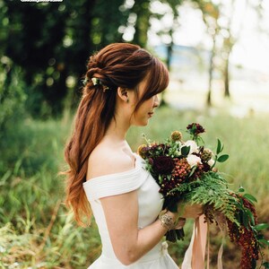 May include: A bride in a white off-the-shoulder wedding dress holds a bouquet of burgundy and white flowers with greenery. Her auburn hair is styled with flowers. The image is set outdoors with a blurred green background.