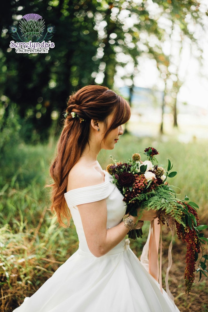 May include: A bride in a white off-the-shoulder wedding dress holds a bouquet of burgundy and white flowers with greenery. Her auburn hair is styled with flowers. The image is set outdoors with a blurred green background.