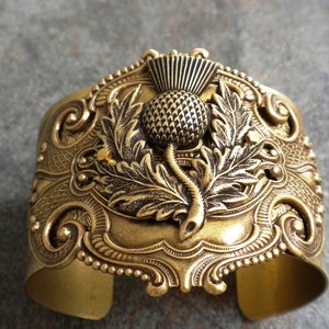 May include: A wide, antique gold-toned cuff bracelet. The bracelet features a detailed thistle design at the center, surrounded by ornate scrollwork and beaded edges. The cuff is made of metal and has a slightly curved shape.