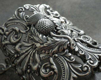 May include: Ornate silver-toned metal bracelet featuring a detailed thistle design. The bracelet showcases intricate scrollwork and leaf patterns, creating a vintage or antique aesthetic. The thistle is the focal point.
