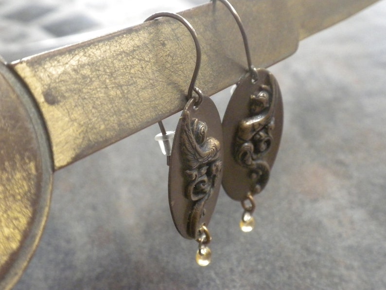 Puede incluir: Un par de pendientes antiguos de color dorado con un dise&ntilde;o detallado de un p&aacute;jaro y motivos florales. Cada pendiente tiene un peque&ntilde;o colgante de piedra preciosa amarilla.