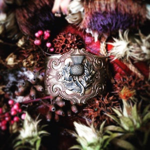 May include: A detailed, antique-style cuff bracelet with a thistle emblem. The metal cuff is a warm bronze color, with the thistle in silver. The background is a mix of dried flowers and berries in shades of red, brown, and green.