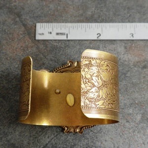May include: A gold-colored cuff bracelet with an ornate floral design. The cuff is approximately 2 inches wide and features detailed leaf patterns and decorative edges. The bracelet has a vintage aesthetic.