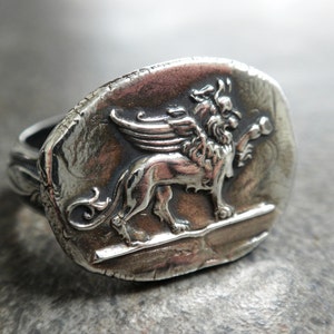 May include: A silver ring with a detailed design of a winged lion. The ring is oval shaped and has a textured surface.