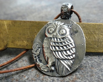 May include: Silver owl pendant necklace with a brown leather cord. The owl is detailed with feathers and has large eyes.