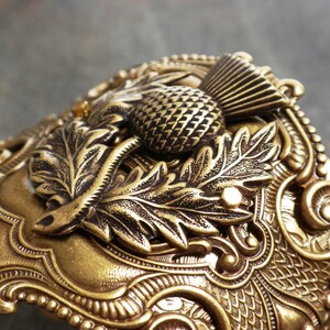 May include: A detailed, antique-style cuff bracelet in a warm, golden hue. The ornate design features a prominent thistle, leaves, and intricate scrollwork. The bracelet has a textured surface and a vintage aesthetic.