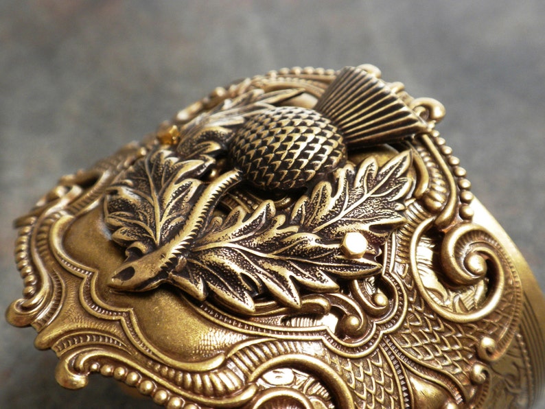 May include: A detailed, antique-style cuff bracelet in a warm, golden hue. The ornate design features a prominent thistle, leaves, and intricate scrollwork. The bracelet has a textured surface and a vintage aesthetic.