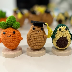 May include: A trio of crocheted food figures: a carrot with green leaves, a potato in a graduation cap, and an avocado. Each is on a small wooden base. The figures have smiling faces and are brightly coloured.