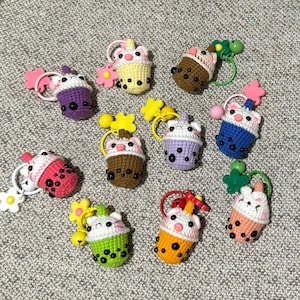 May include: A selection of crocheted bubble tea keyrings in various colours. Each keyring features a cup with a cute animal face, decorated with a flower or other details. The keyrings are in shades of pink, purple, yellow, and green.