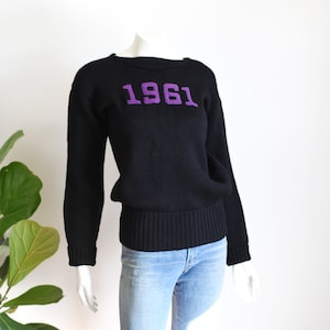May include: Black knitted jumper with the number "1961" in purple lettering across the chest. The jumper has a ribbed hem and cuffs. The jumper is displayed on a mannequin wearing light blue jeans.