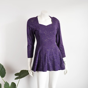 May include: A purple dress with a square neckline and three-quarter sleeves. The dress has a fitted bodice and a flared skirt. Gold flecks are scattered across the fabric. The dress is displayed on a mannequin.