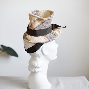 Könnte beinhalten: Ein Hut im Vintage-Stil mit einem einzigartigen Design. Der Hut hat einen cremefarbenen Stoff mit einem Karomuster in Grau-, Rosa- und Brauntönen. Ein dunkelbraunes Band umschließt den Hut. Der Hut wird auf einem weißen Schaufensterkopf präsentiert.