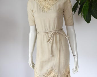 Puede incluir: Vestido de lino beige con mangas cortas y cuello barco. El vestido presenta intrincados detalles de encaje alrededor del escote y el dobladillo, y un cinturón a juego ciñe la cintura. El vestido se exhibe en un maniquí.