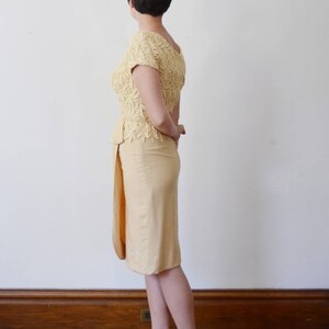 Puede incluir: Una mujer con un vestido de encaje amarillo con una falda que le llega a las rodillas. Est&aacute; de pie sobre un suelo de madera con una pared blanca detr&aacute;s de ella.